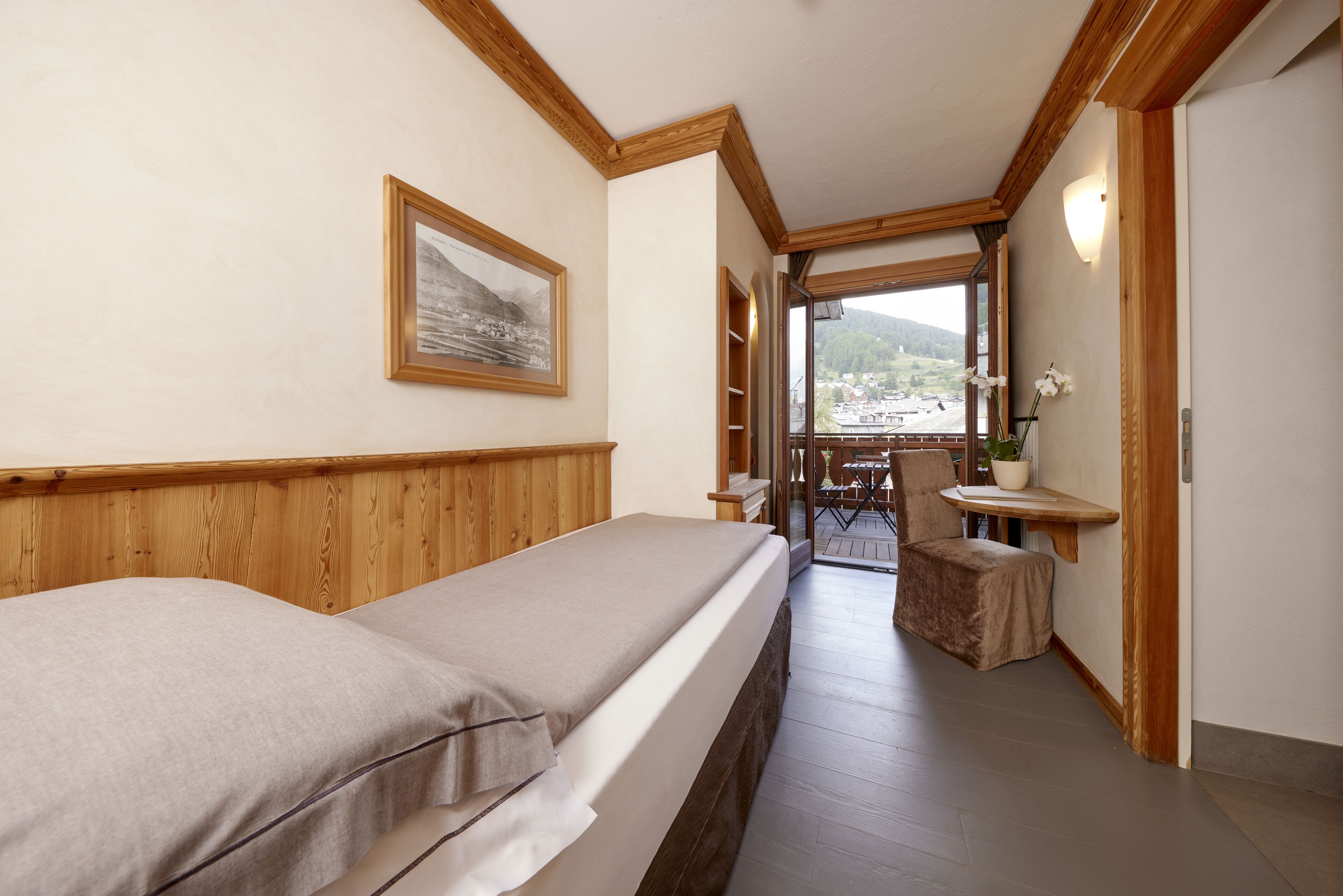 A hotel room with two beds, a wooden headboard, a framed picture on the wall, a small table with a potted plant, and a view of a balcony with outdoor furniture.