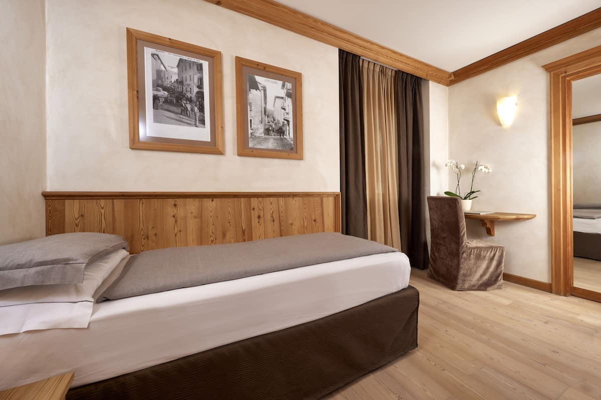 Una cama bien hecha con cabecera de madera, dos fotografías en blanco y negro enmarcadas en la pared, una mesita de noche con un jarrón de flores blancas y un espejo grande a la derecha.