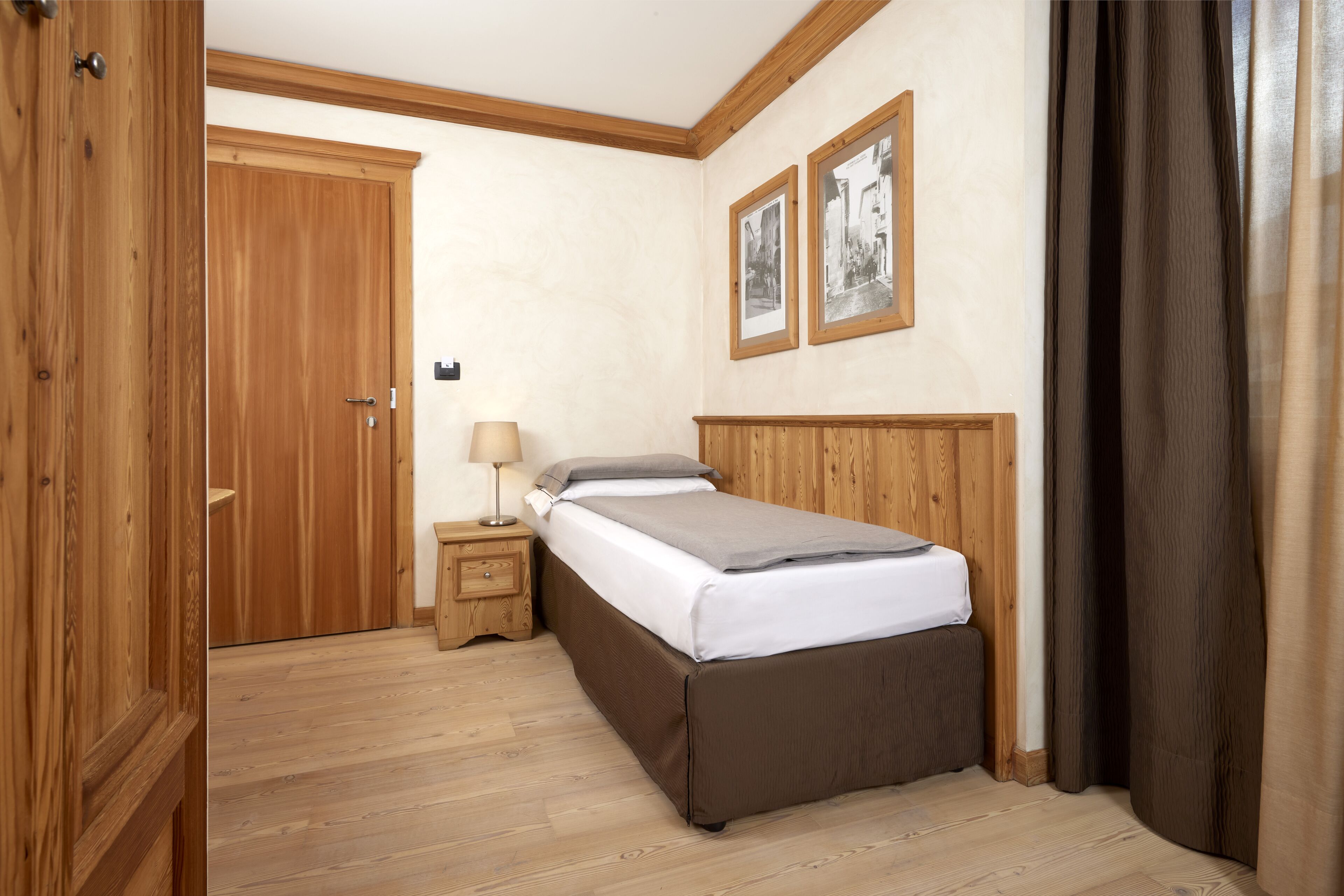 Une chambre avec une armoire en bois, une table de chevet avec une lampe, un lit recouvert d’une couverture grise et deux tableaux encadrés accrochés au mur.