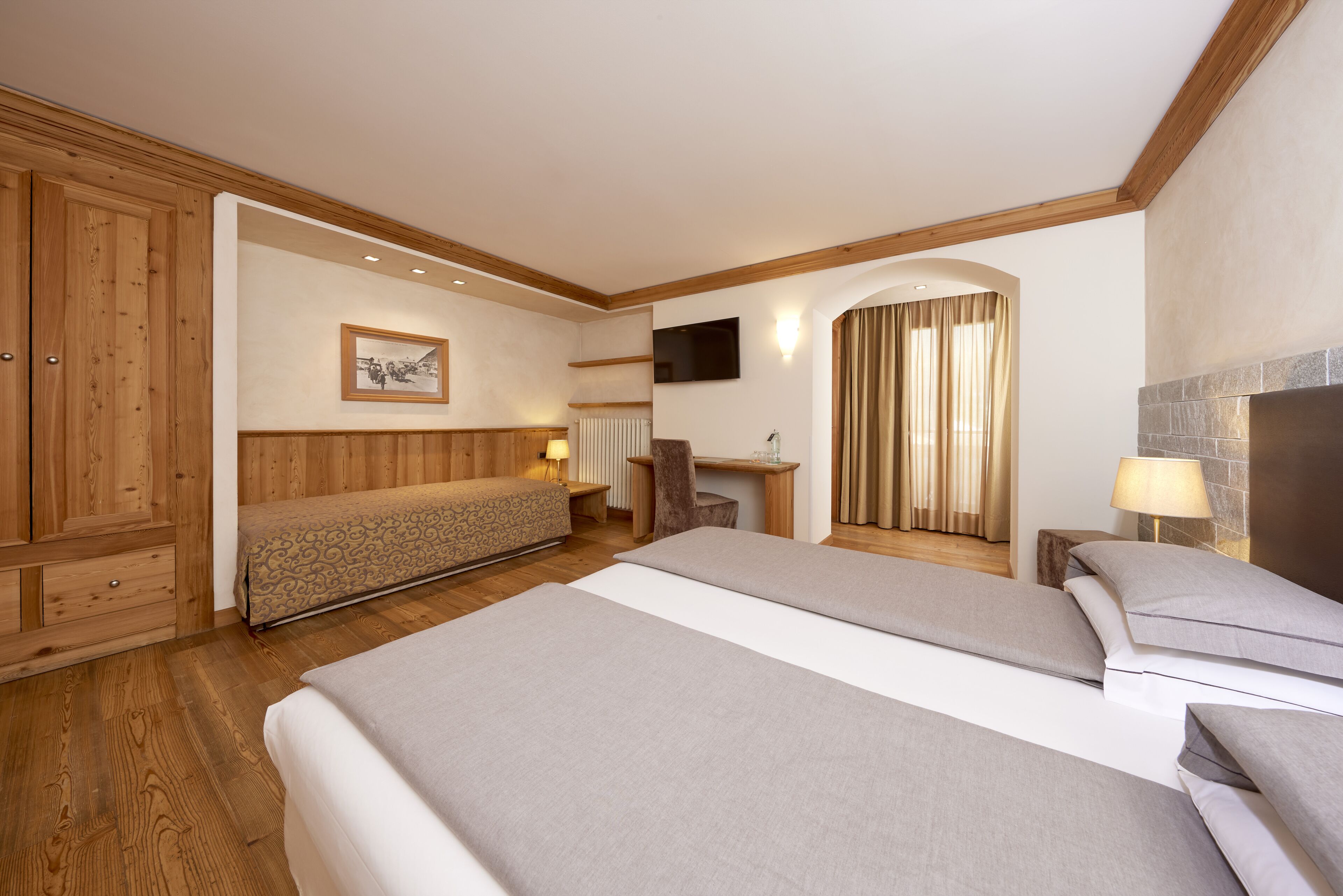 A hotel room with a large bed, a wooden wardrobe, a desk, a chair, a television, and a fireplace.