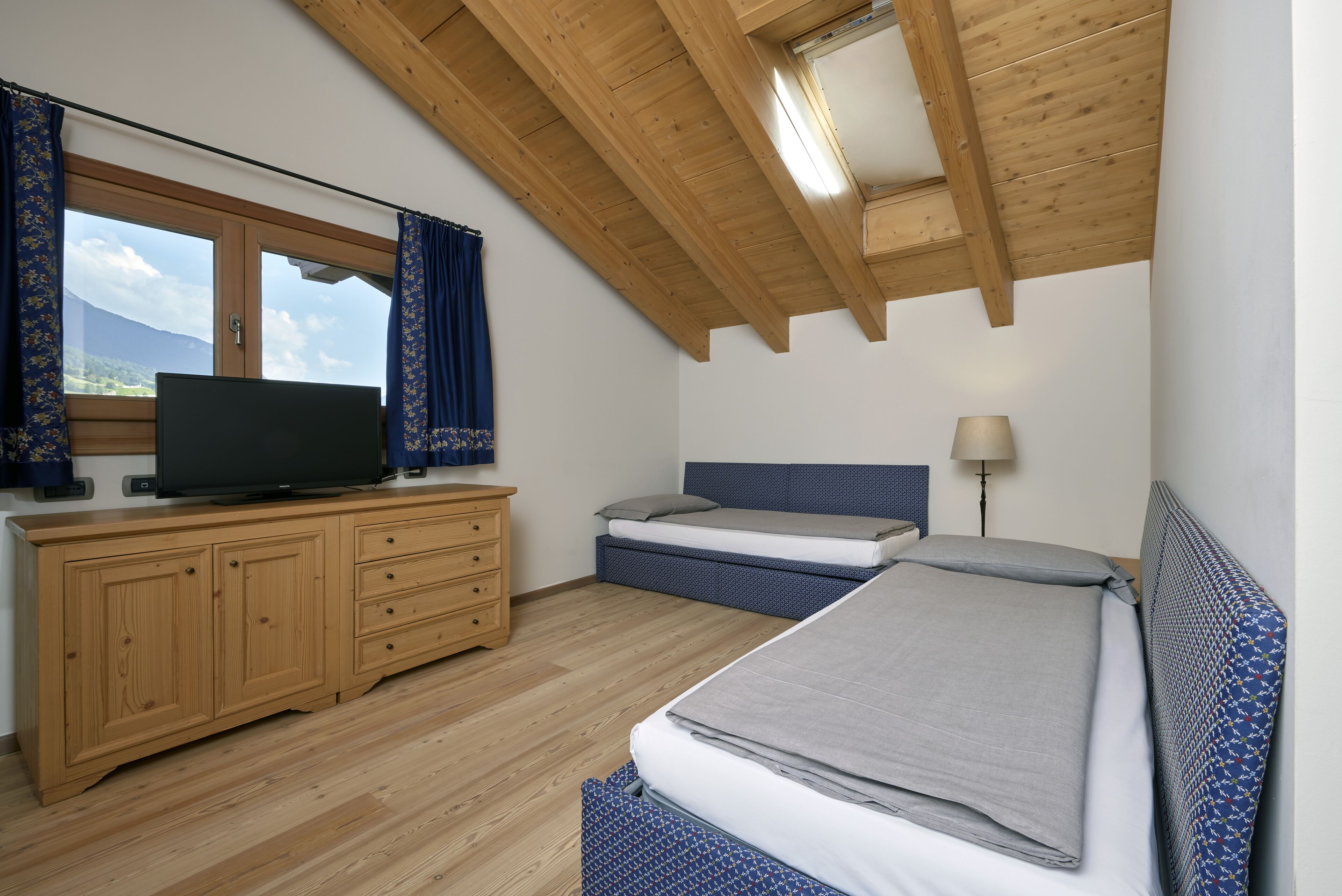 A bedroom with a wooden ceiling, a TV on a wooden dresser, a bed with a blue headboard, and a window with curtains.