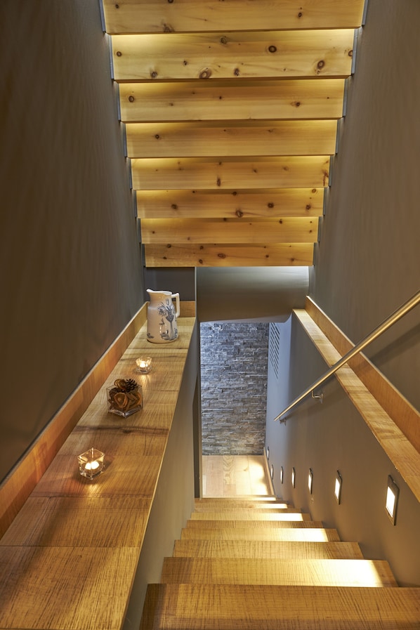 Eine Holztreppe mit einer Steinwand am unteren Ende, ein kleiner Tisch mit einer Vase und Kerze sowie eine Holzbalkendecke.