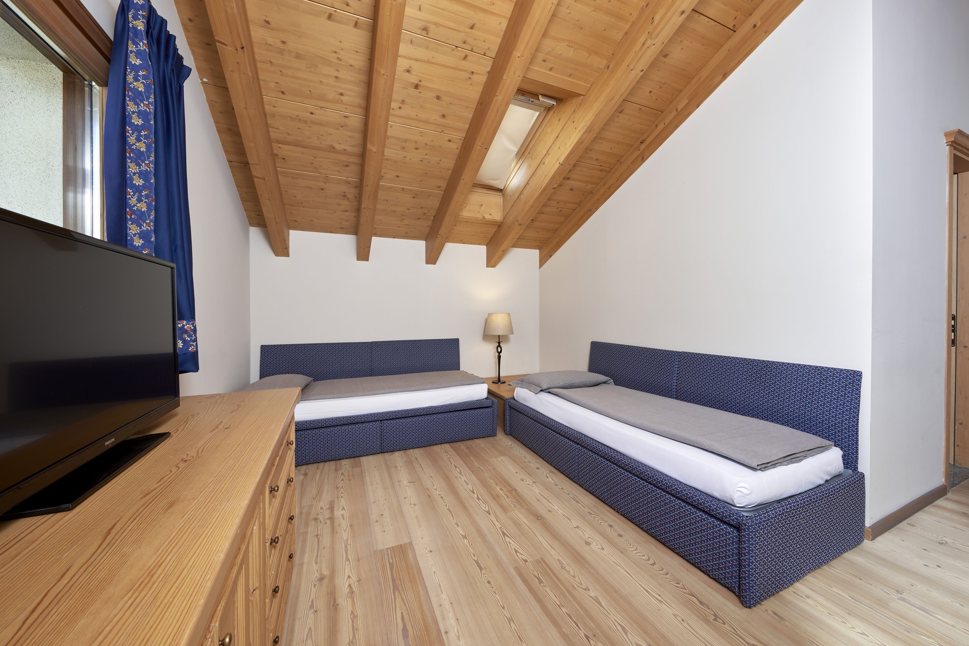 Una habitación con techo de madera, dos camas con cabeceros azules, un televisor sobre una mesa de madera y una lámpara en una mesita de noche.