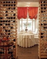 Una bodega con estantes llenos de botellas de vino, una mesa redonda dispuesta para cenar y una ventana con cortinas.