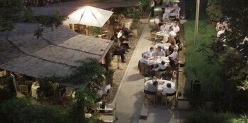 Un área de comedor al aire libre con mesas y sillas, rodeada de follaje y un edificio rústico con tejado de paja.