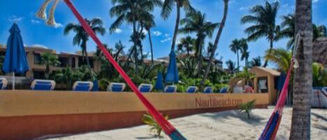 A beach with a hammock, palm trees, and a building with a sign that reads "Nautibeach.com".