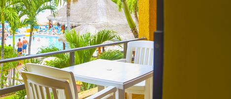 Un balcón con sillas y una mesa blancos, con vista a una piscina con tumbonas y palmeras.
