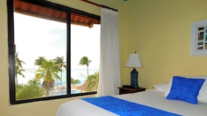 Une chambre d’hôtel avec un lit, une table de chevet, une lampe, une fenêtre donnant sur la plage et les palmiers, et un tableau accroché au mur.