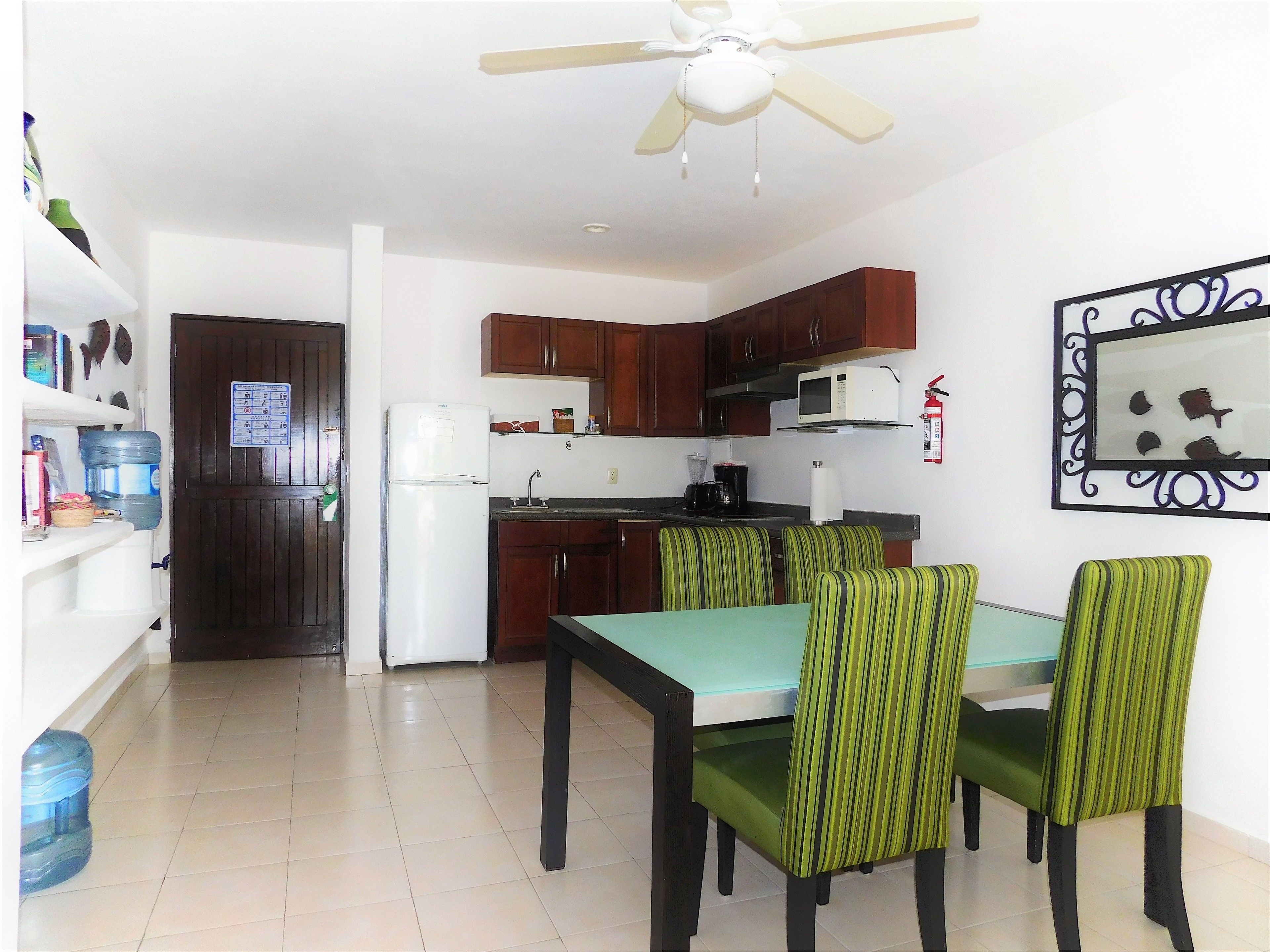 A compact kitchen with wooden cabinets, a white refrigerator, a microwave, and a coffee maker. There is a dining table with green-striped chairs.