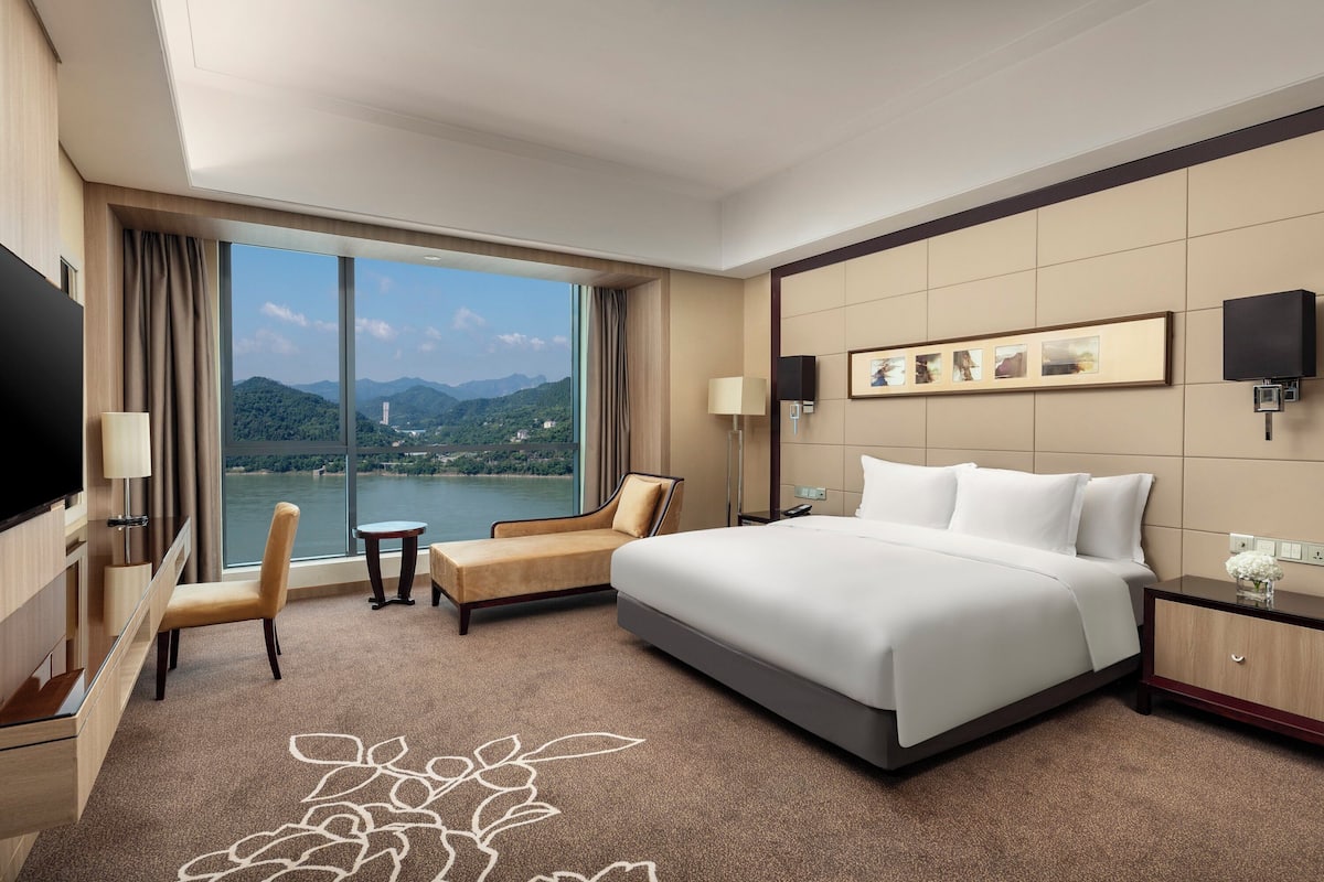 Habitación de hotel con una cama grande, un escritorio, una silla, un televisor y vistas a un lago y montañas.