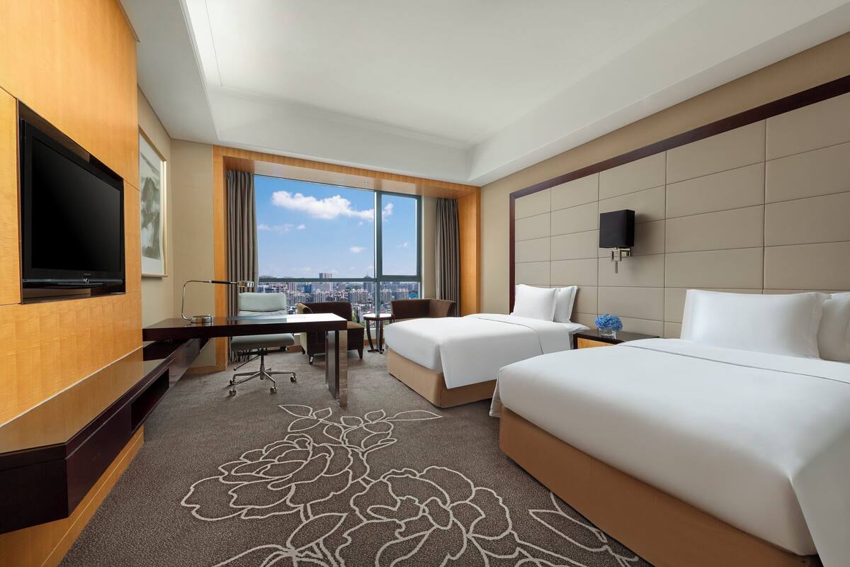 Habitación de hotel con dos camas, un escritorio, televisión y un amplio ventanal con vistas a la ciudad.
