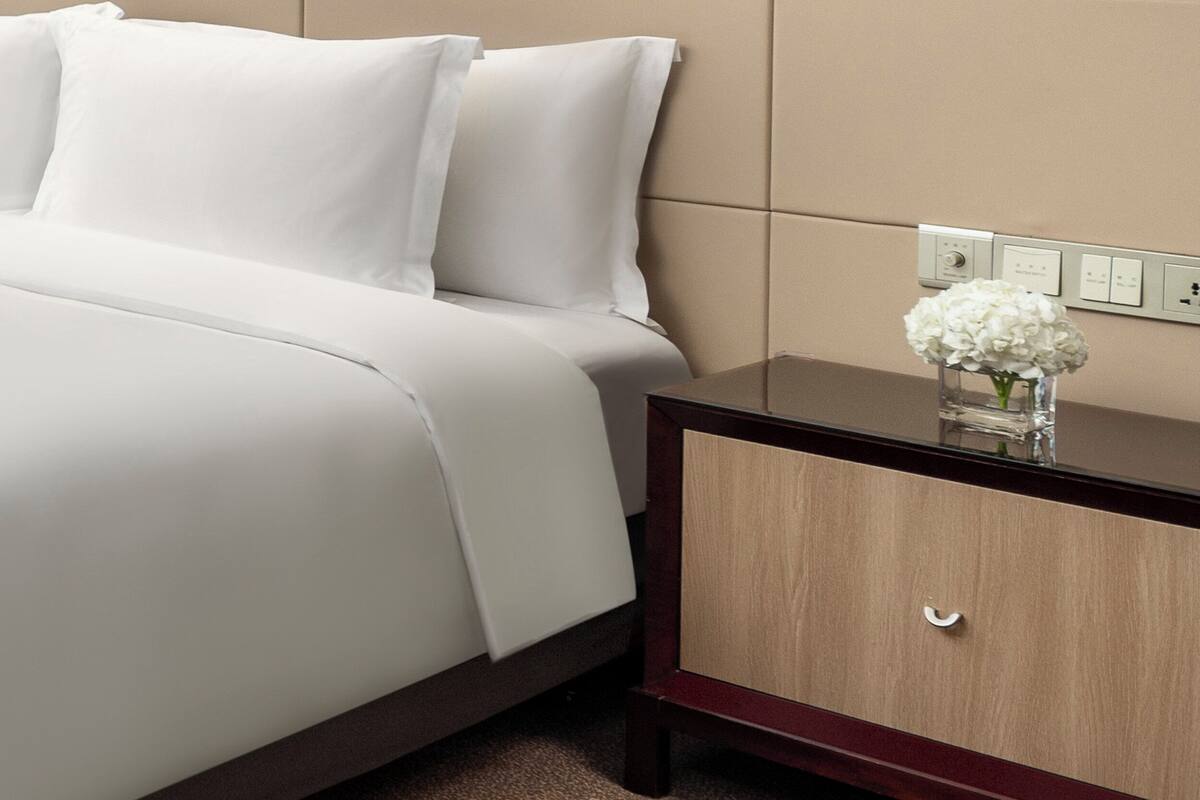 Una cama bien hecha con sábanas blancas, una mesita de noche de madera con un cajón y un florero con flores blancas.