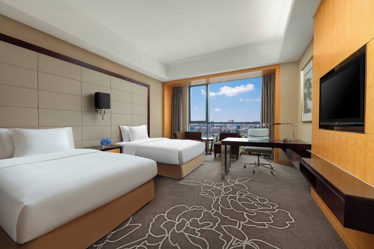 Habitación de hotel con dos camas, un escritorio, una silla, un televisor y un ventanal grande con vistas a la ciudad.