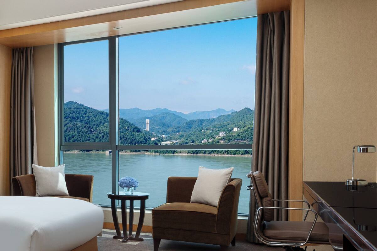 Habitación de hotel con un amplio ventanal que ofrece vistas a un río y montañas. Cuenta con una cama, dos sillones, una mesita con un florero y un escritorio con una lámpara.