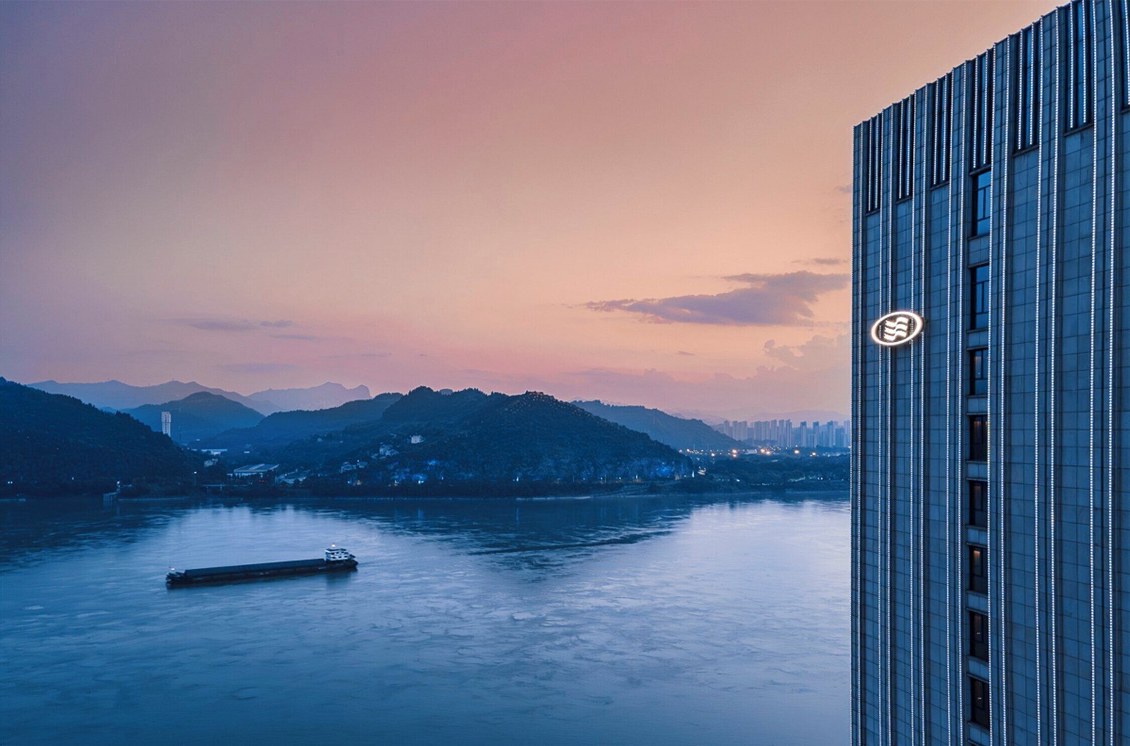 Um edifício moderno com um logotipo em destaque, com vista para um rio calmo e uma cordilheira distante, além do horizonte da cidade.