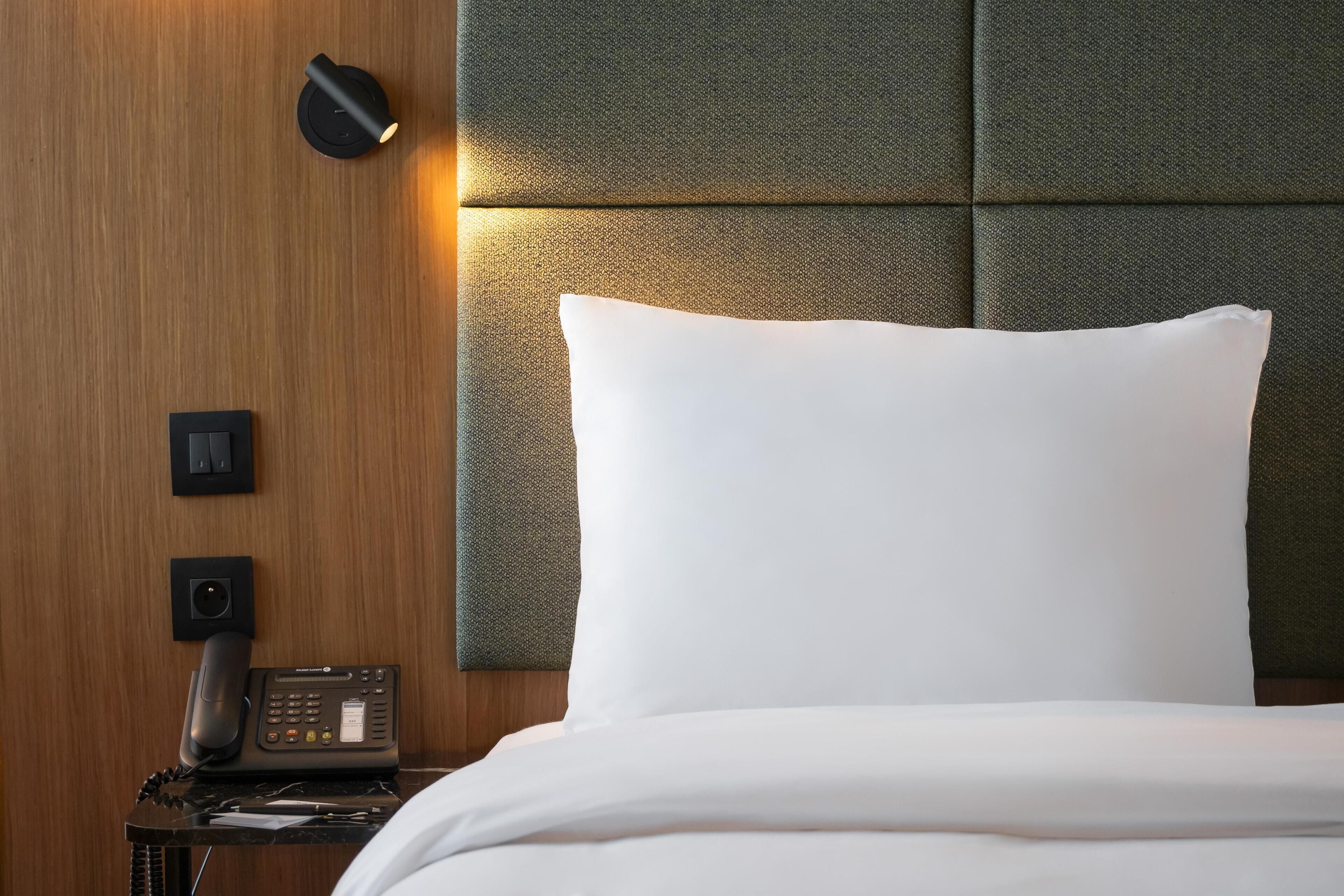 A hotel room with a bed, a bedside telephone, and a wall-mounted light fixture.