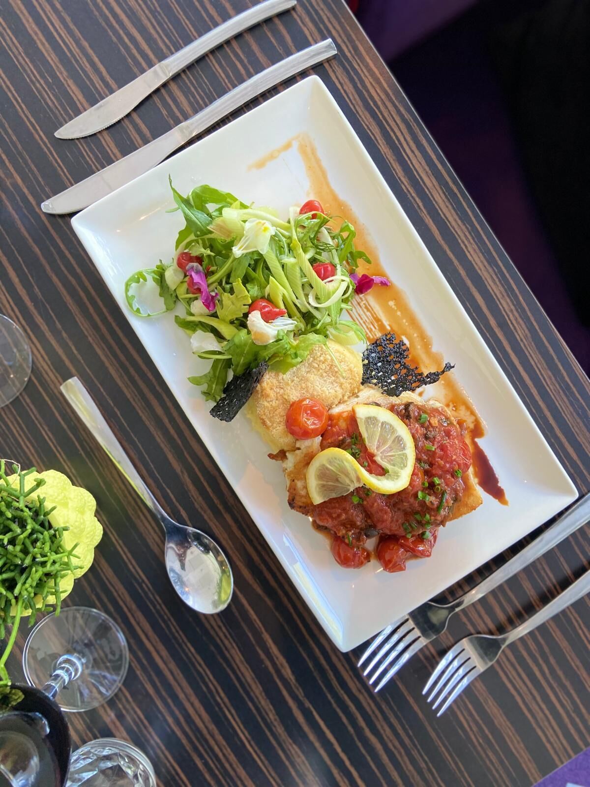A rectangular white plate with a fish fillet topped with lemon slices and cherry tomatoes, accompanied by a side salad and a small piece of dark, crispy garnish.