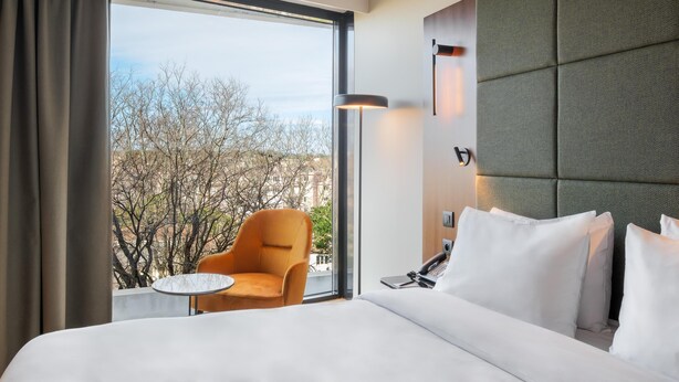 A hotel room with a large bed, a chair, a small table, and a view of trees and buildings. - Radisson Blu Hotel, Montpellier (Montpellier)