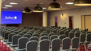 A conference room with rows of chairs, a large screen displaying Radisson BLU logo, and pendant lighting.