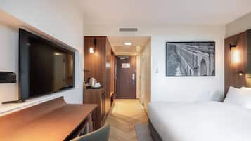 Une chambre d’hôtel avec un lit, un bureau doté d’un téléviseur à écran plat, une chaise, un plancher en bois et un tableau encadré au mur.
