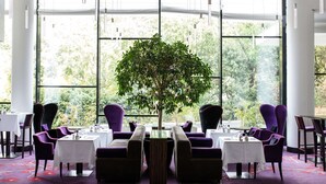 A dining area with tables and chairs, a central plant, and large windows with a view of trees. - Radisson Blu Hotel, Montpellier (Montpellier)