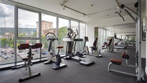 Un gimnasio con diferentes máquinas de ejercicio, grandes ventanales y vistas a edificios y árboles.
