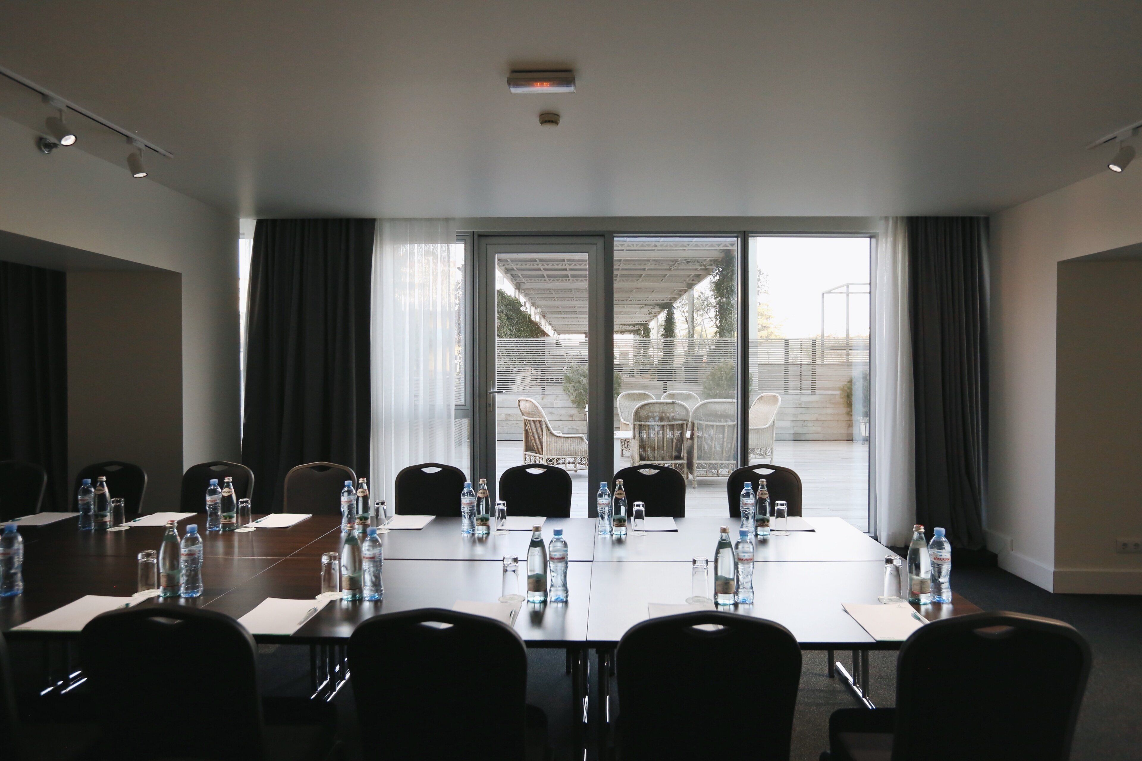 A conference room with a long table, chairs, water bottles, and a view through glass doors to an outdoor area with chairs and a table.