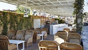 Un área de estar al aire libre con sillas y mesas de mimbre, un bar con mostrador, y un fondo de follaje y edificios.
