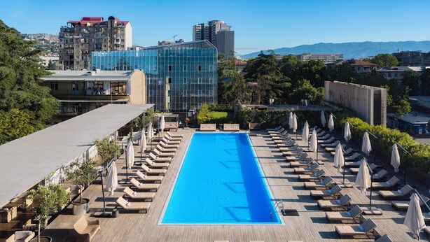 A rooftop pool with lounge chairs and umbrellas, surrounded by buildings and trees.