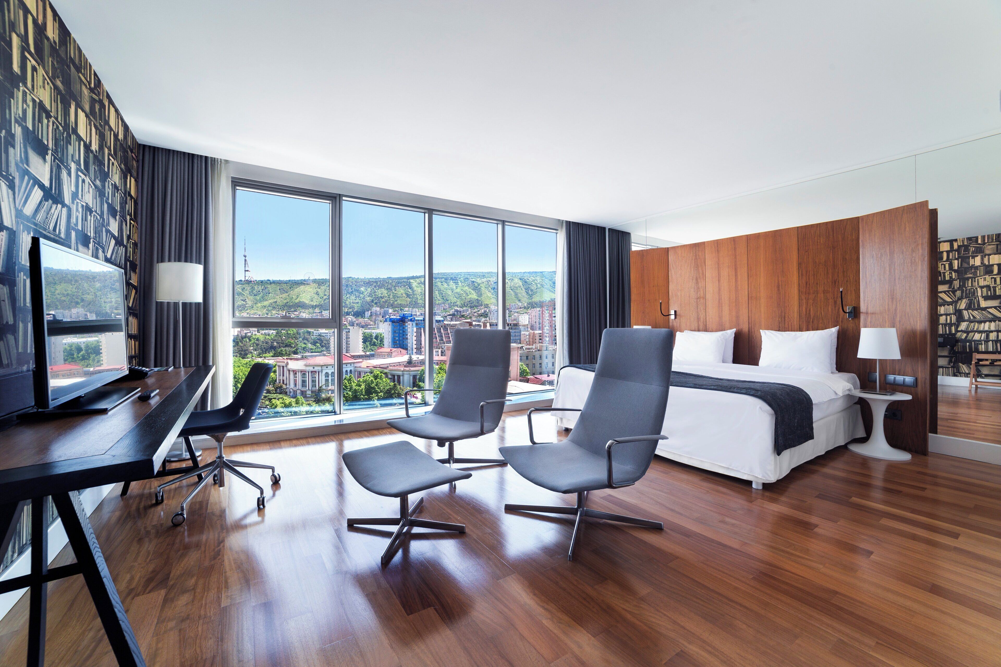 A modern hotel room with a large window, a desk with a chair, a sofa, a bed, and a bookshelf.