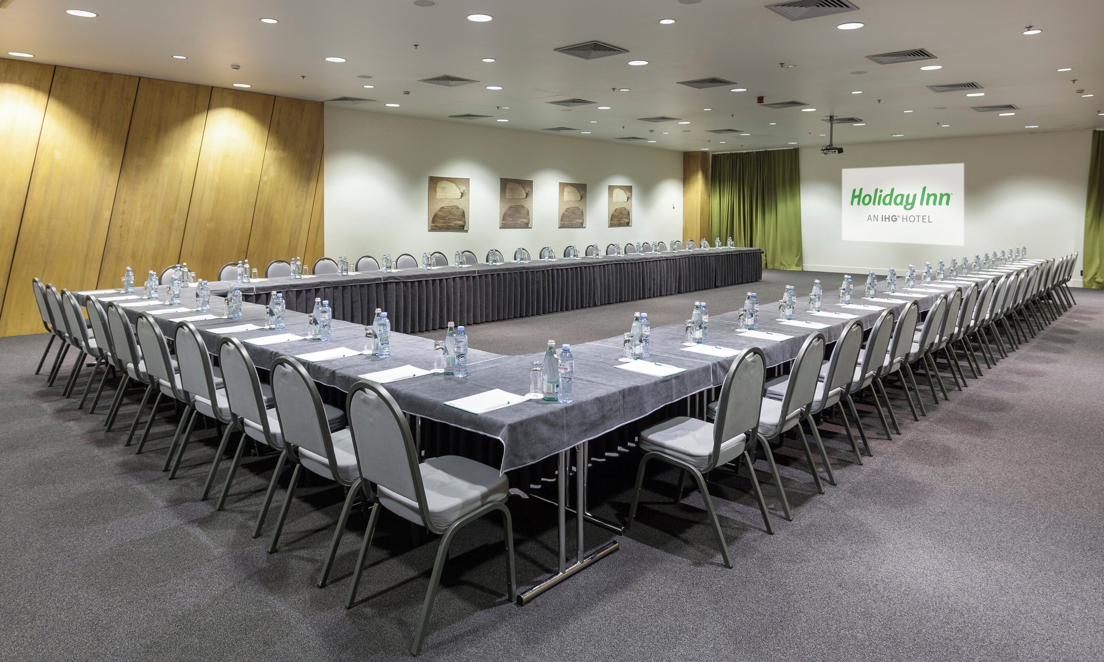 Un salón de conferencias con una larga mesa, sillas, botellas de agua y una pantalla proyectora mostrando 'Holiday Inn'.