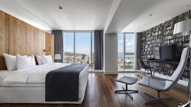 Una habitación de hotel moderna con una cama grande, un escritorio con silla, una estantería y vistas a la ciudad.