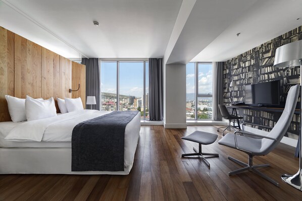 A modern hotel room with a large bed, a desk with a chair, a bookshelf, and a city view. - Holiday Inn Tbilisi by IHG (Tbilisi)