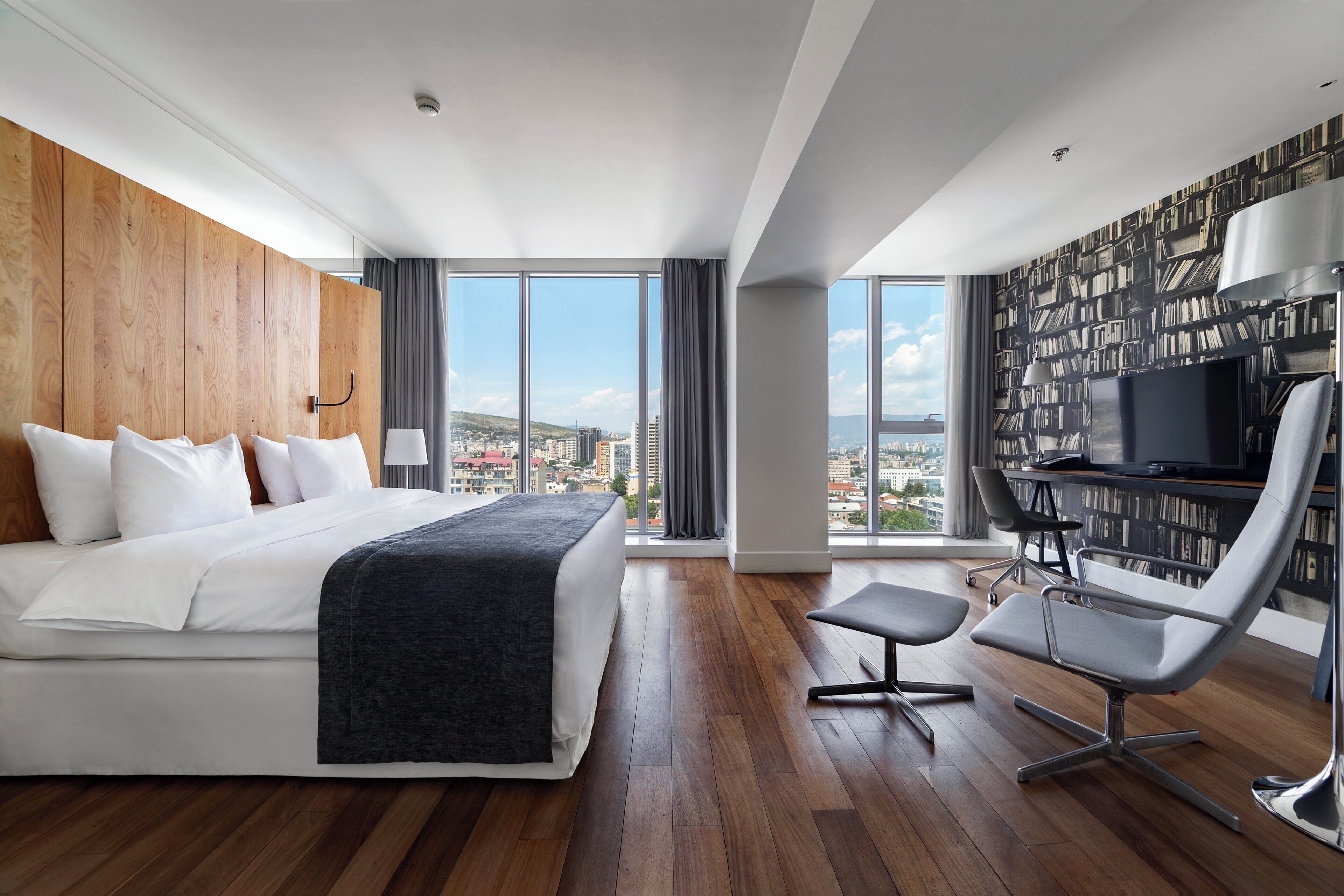 Habitación de hotel moderna con una cama grande, un escritorio con silla, una estantería y vista a la ciudad.