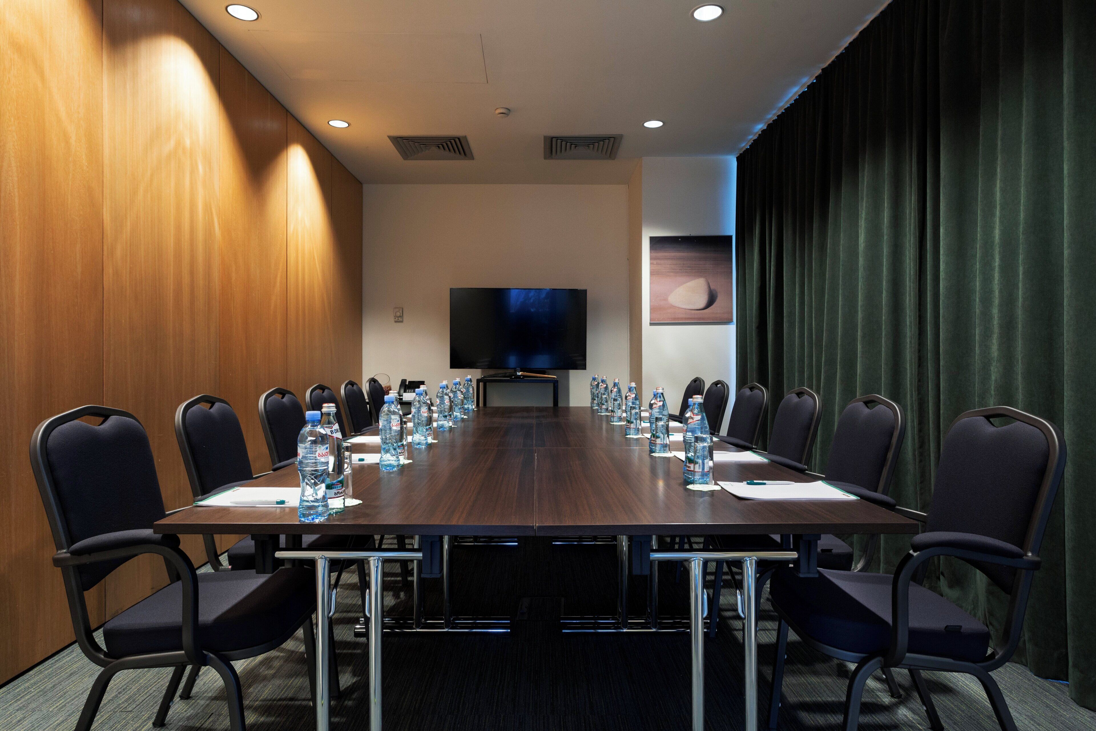Een vergaderzaal met een lange tafel, stoelen, waterflesjes, een flatscreen televisie en een schilderij aan de muur.