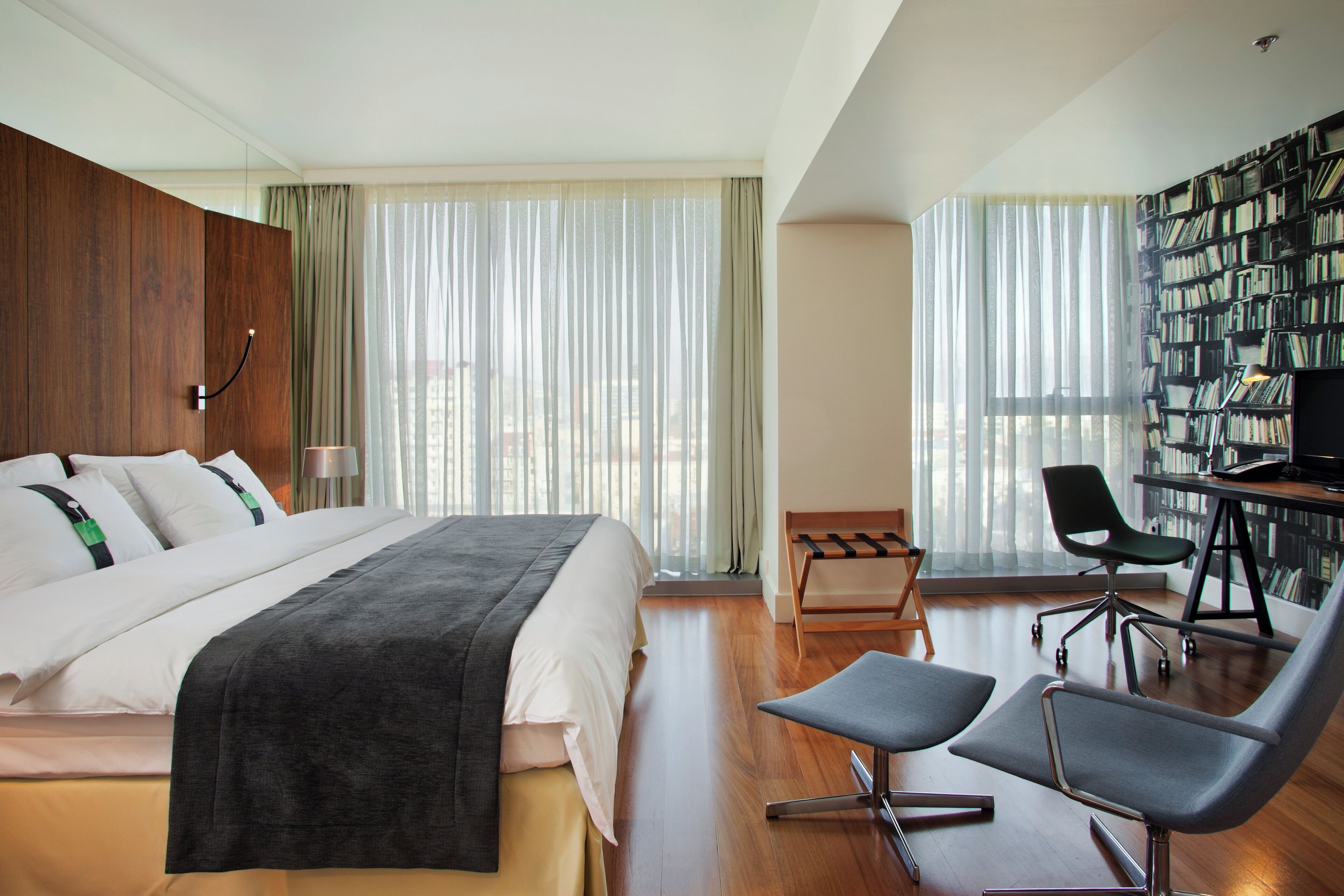 Una habitación de hotel moderna con una cama grande, un escritorio con silla, una estantería y ventanas amplias con cortinas ligeras.