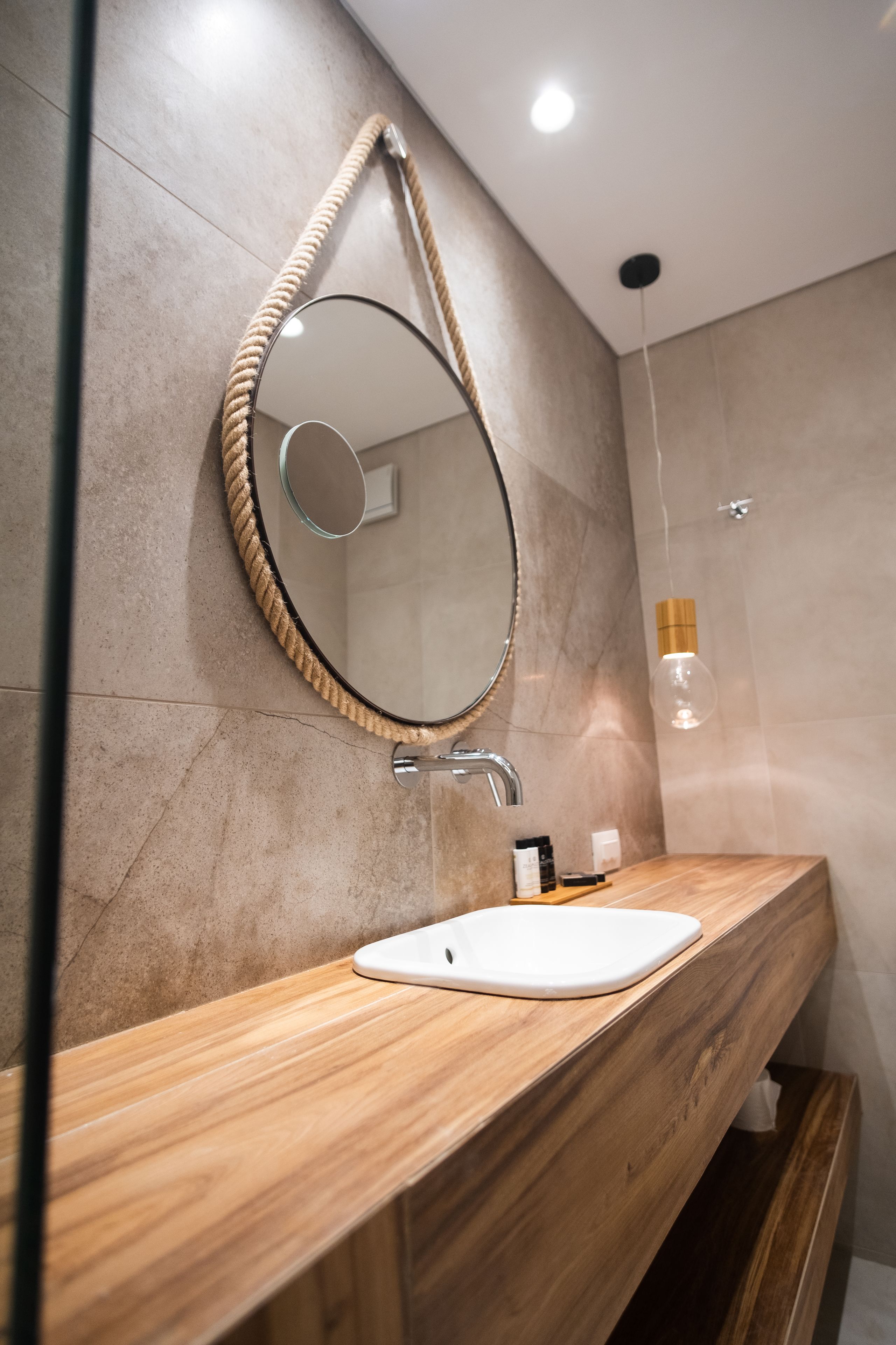 A bathroom with a wooden vanity, a round mirror with a rope frame, a white sink, and a shower area with a wooden door.