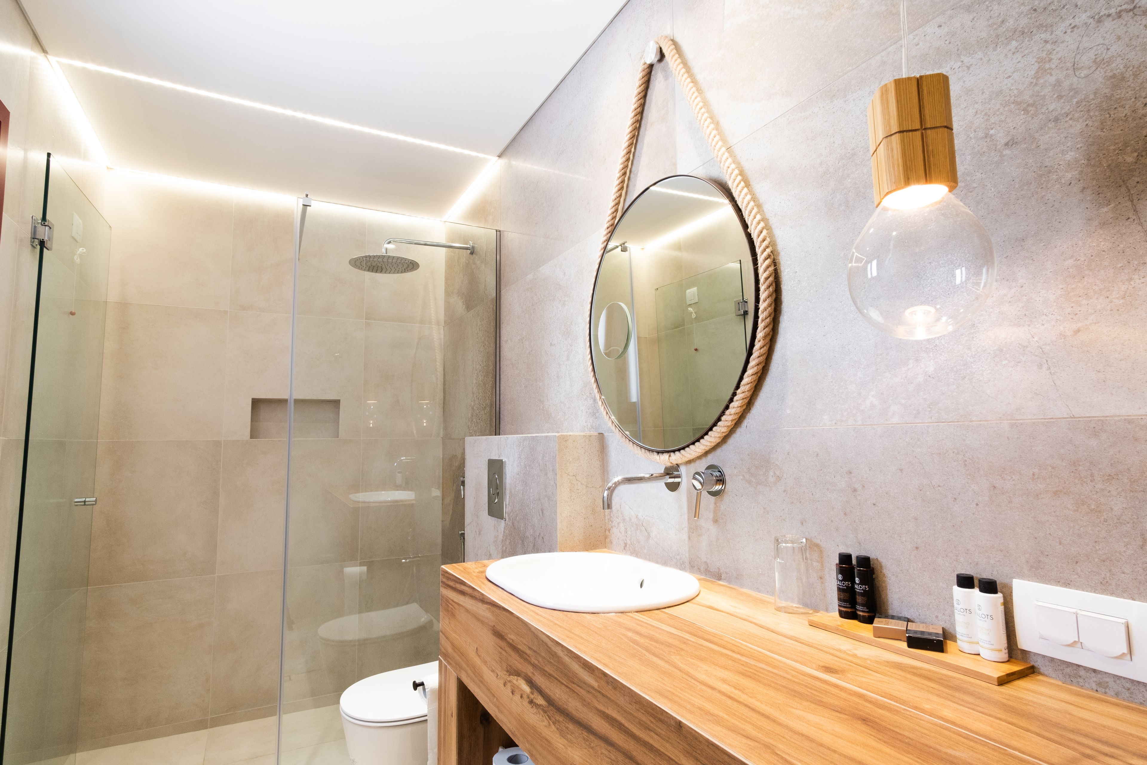 Un baño moderno con ducha de vidrio, tocador de madera con lavamanos blanco y espejo con asa de cuerda.
