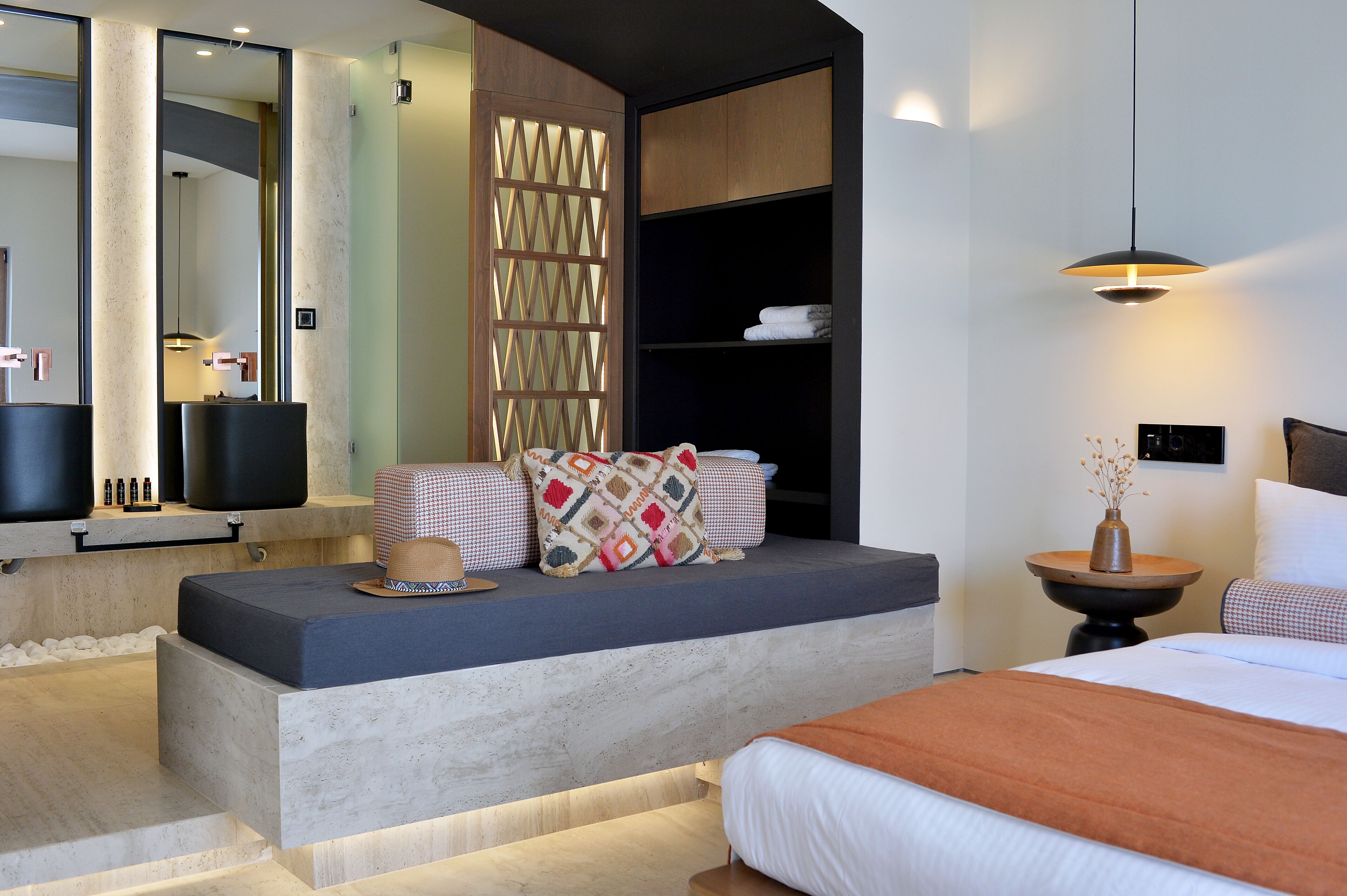 Una habitación de hotel moderna con una cama grande, un área de descanso con cojines, una mesita con un jarrón y un espejo amplio.