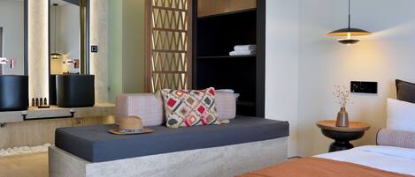 Una habitación de hotel moderna con una cama grande, un área de descanso con cojines, una mesita con un jarrón y un espejo amplio.
