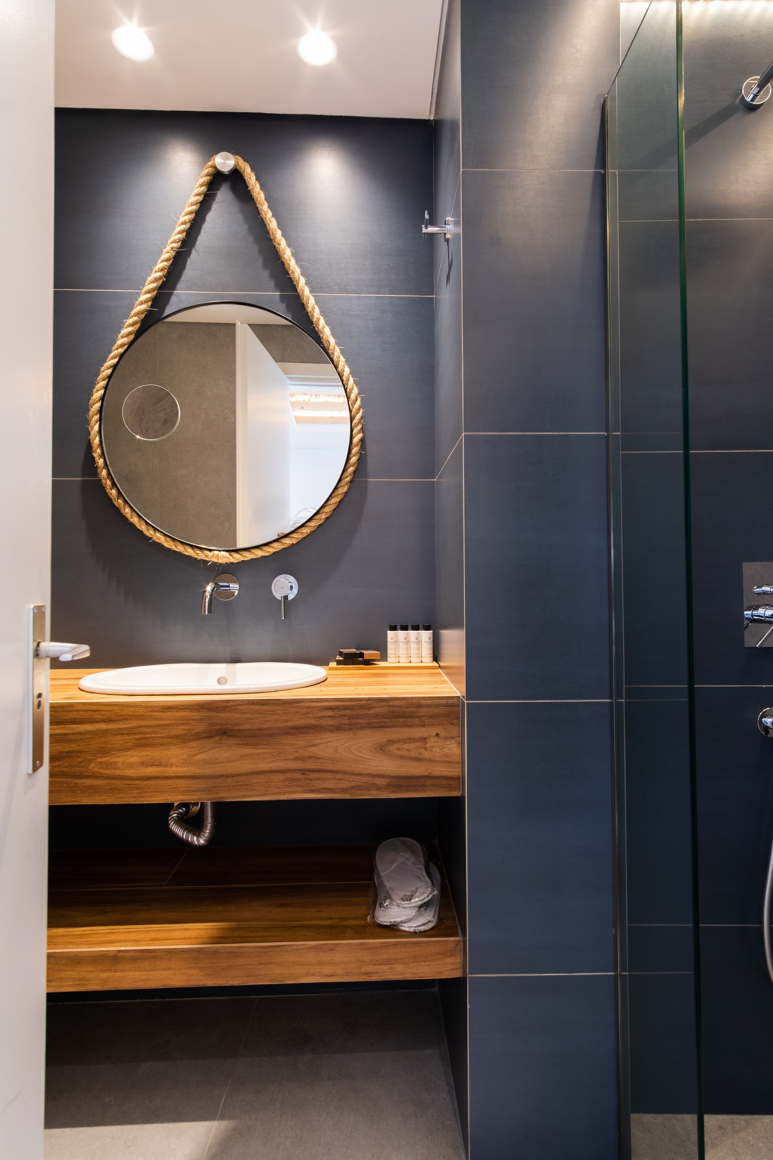 A modern bathroom with a wooden vanity, a round mirror with a rope frame, and a glass shower enclosure.