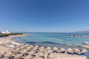 Uma praia com guarda-sóis de palha, espreguiçadeiras e vista clara para o mar e barcos.