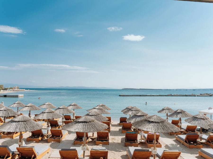 Vista de la playa con sillones de madera y sombrillas de paja, cielo despejado y mar tranquilo.