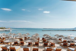Blick auf einen Strand mit Holzliegen und Reetdachschirmen, einem klaren Himmel und ruhiger See.
