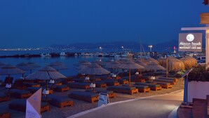 A beachside lounge area with sun loungers and thatched umbrellas, a sign for Aktaion Beach Boutique Hotel, and a view of the marina and city lights in the background.