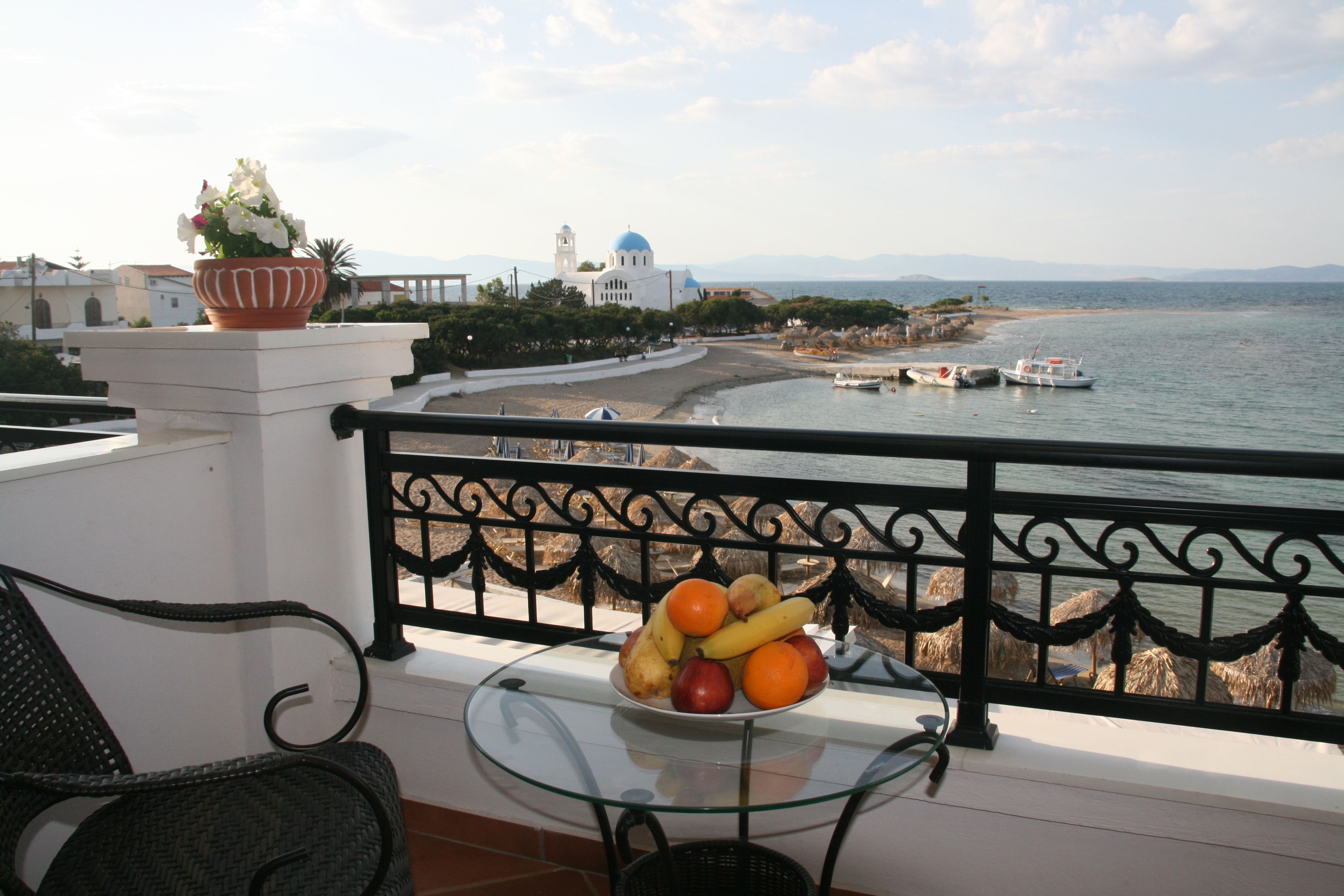 A balcony with a glass table holding fruit, overlooking a beach with boats and a church with a blue dome.