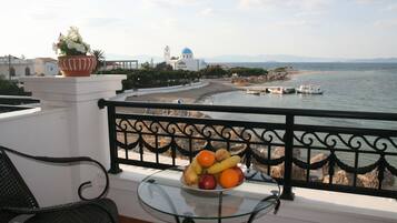 A balcony with a glass table holding fruit, overlooking a beach with boats and a church with a blue dome.