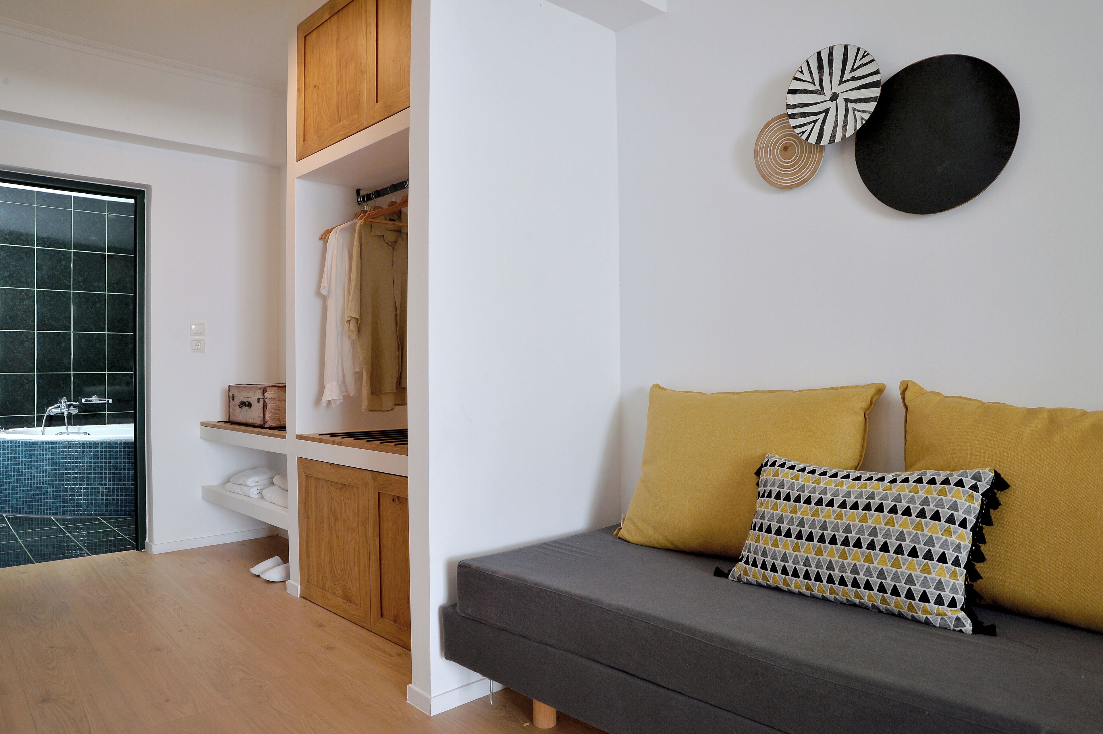 Een moderne woonkamer met een grijze bank, mosterdgele kussens en een geborduurd kussen. Een houten garderobe en een wit boekenplank met opgevouwen kleding zijn zichtbaar.