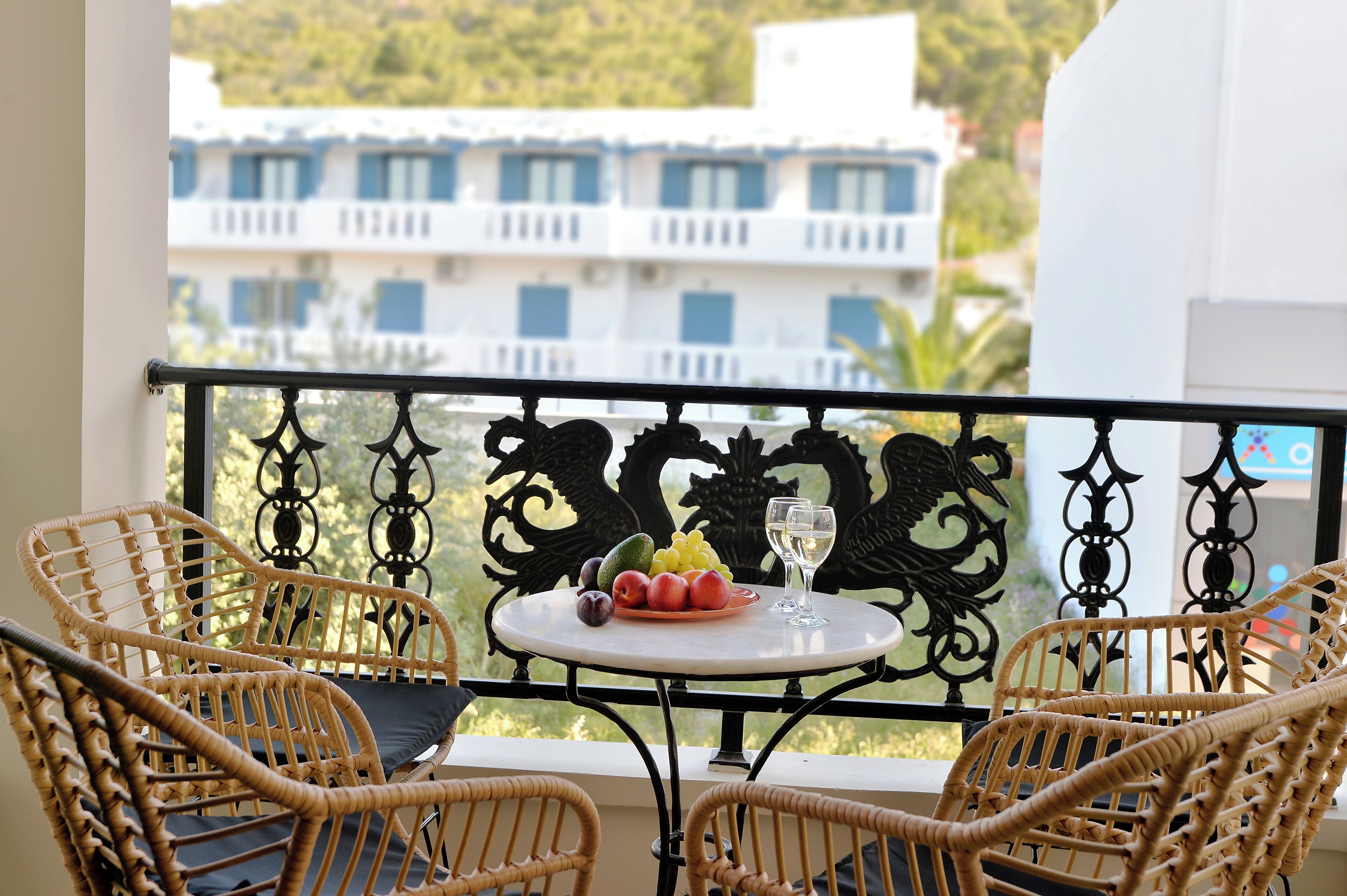 Un balcon avec des fauteuils en osier, une table garnie de fruits et un verre de vin, et une vue sur un immeuble aux volets bleus.