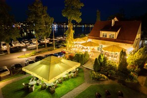 A well-lit outdoor dining area with umbrellas, a lawn, and a view of the water and boats in the background. - Hotel Amax (Mikolajki)